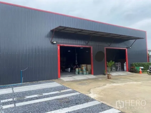 Warehouse for sale in Khlong Sam Wa, Bangkok - Close view of corrugated steel warehouse facade with red trim, canopy and large loading doors beside open parking area.