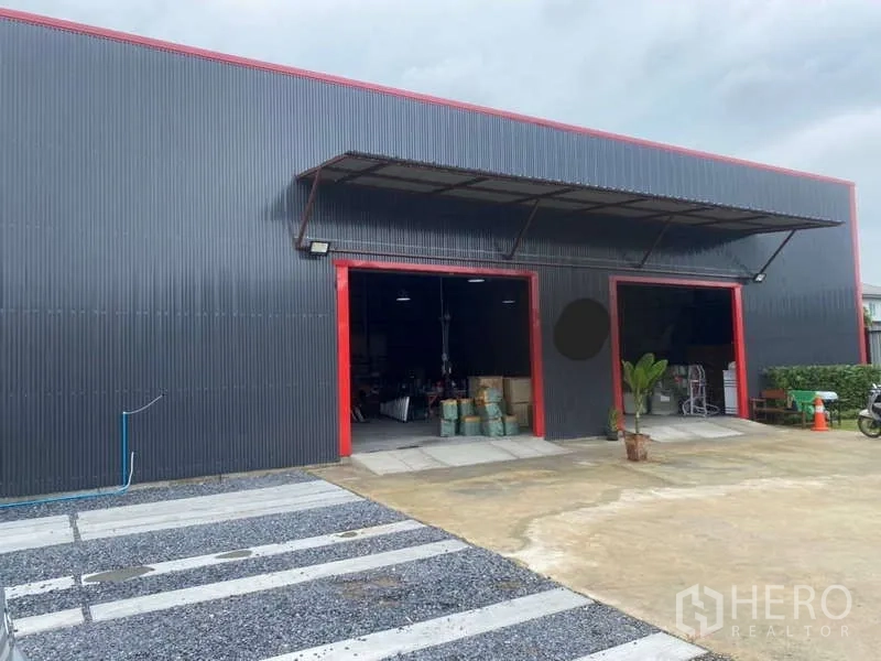 Warehouse for sale in Khlong Sam Wa, Bangkok - Close view of corrugated steel warehouse facade with red trim, canopy and large loading doors beside open parking area.