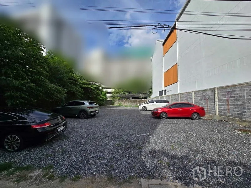 带办公室的家 出租在Chatuchak, Bangkok - 乍都节曼谷低层建筑旁的碎石停车区，停放多辆汽车并有绿树环绕。