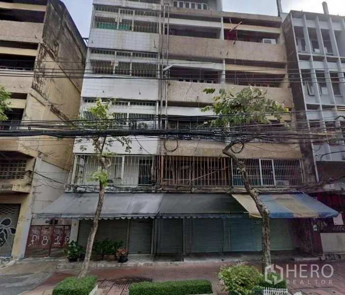 Shophouse for rent in Pom Prap Sattru Phai, Bangkok - Multi-storey shophouse facade with closed roller shutters and balconies on a tree-lined Bangkok street.