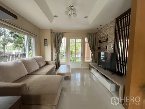 บ้าน ให้เช่าในLat Krabang, Bangkok - ห้องนั่งเล่นสว่าง โซฟารูปตัวแอล ผนังทีวี และหน้าต่างบานใหญ่ติดสวน