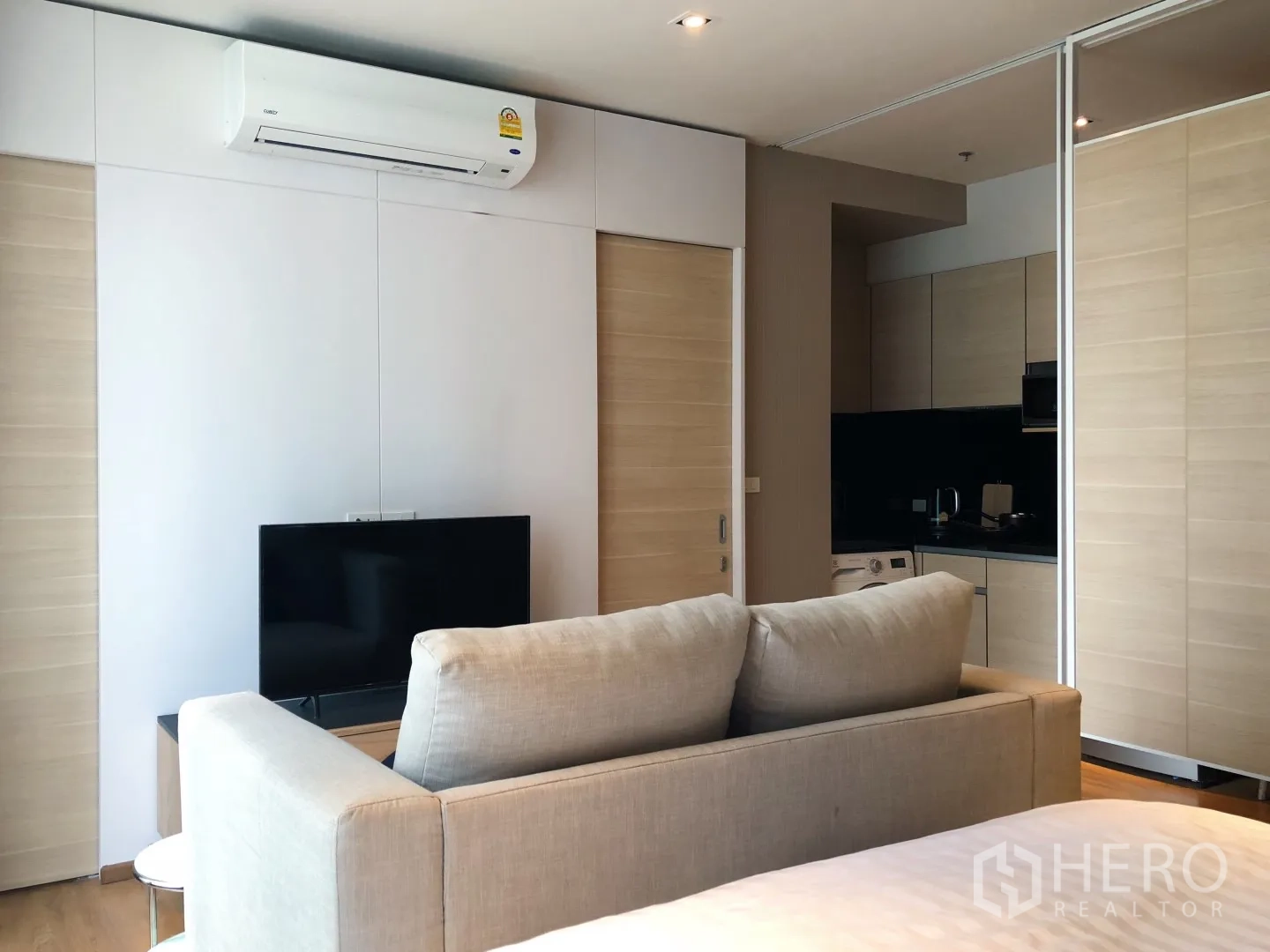 Condominium for rent in Khlong Toei, Bangkok - Open-plan living area with beige sofa, TV unit, air conditioner and sliding doors to the kitchen.