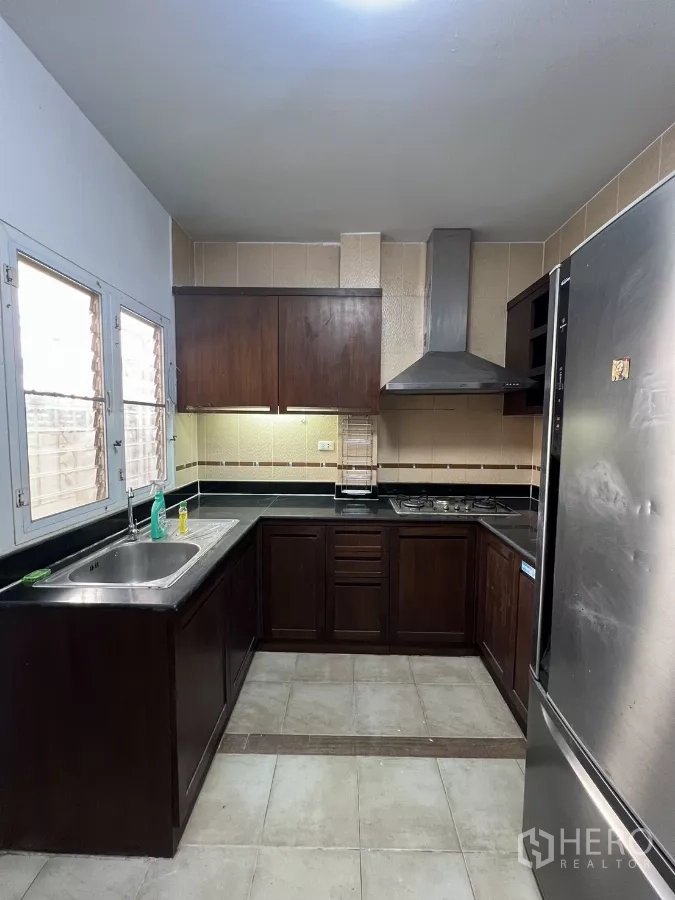 Detached House for rent in Bang Kapi, Bangkok - U-shaped kitchen with dark wood cabinets, sink, windows and gas hob with hood.