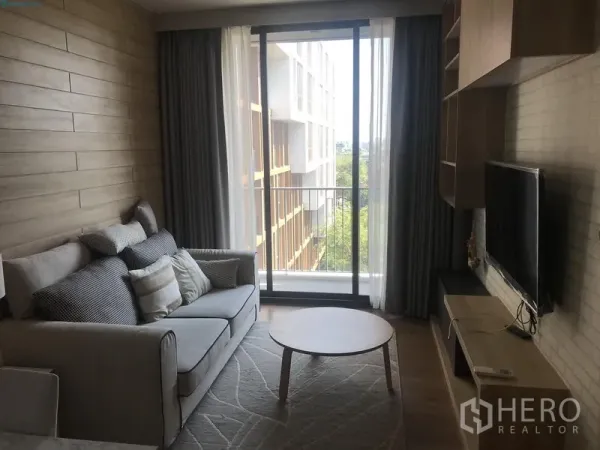 Condominium for rent in Watthana, Bangkok - Cozy living area with TV console and sofa opening to a balcony through floor‑to‑ceiling sliding glass, with green views.