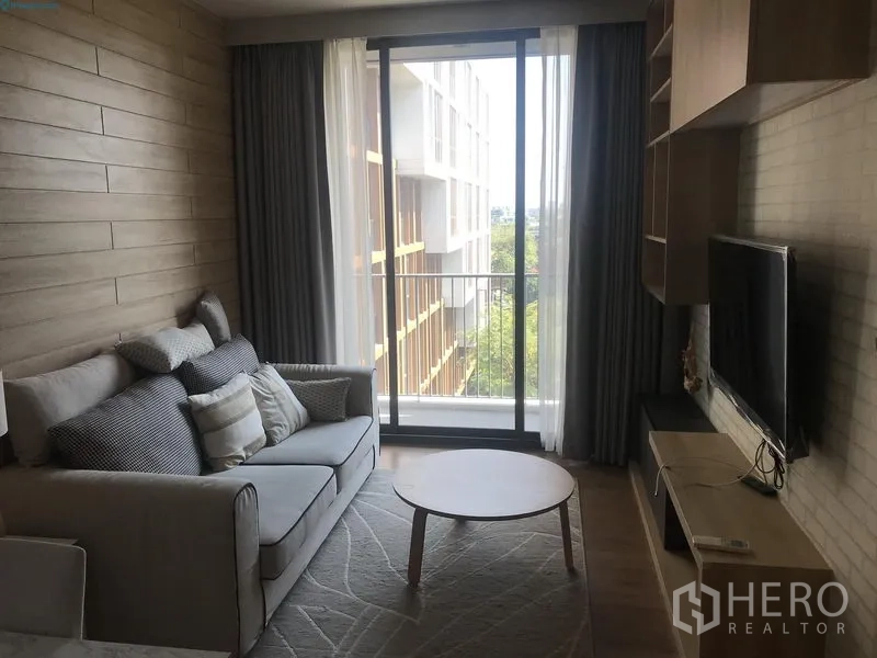 Condominium for rent in Watthana, Bangkok - Cozy living area with TV console and sofa opening to a balcony through floor‑to‑ceiling sliding glass, with green views.
