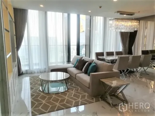 Condominium for rent in Pathum Wan, Bangkok - Sunlit living room with L-shaped sofa, rug, round coffee table and chandelier dining set.