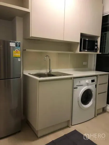 Condominium for rent or sale in Huai Khwang, Bangkok - Compact kitchenette with white cabinetry, sink, washing machine, microwave, and refrigerator.