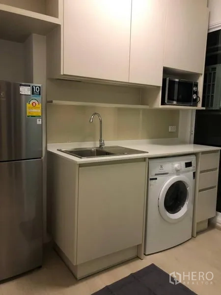 Condominium for rent or sale in Huai Khwang, Bangkok - Compact kitchenette with white cabinetry, sink, washing machine, microwave, and refrigerator.
