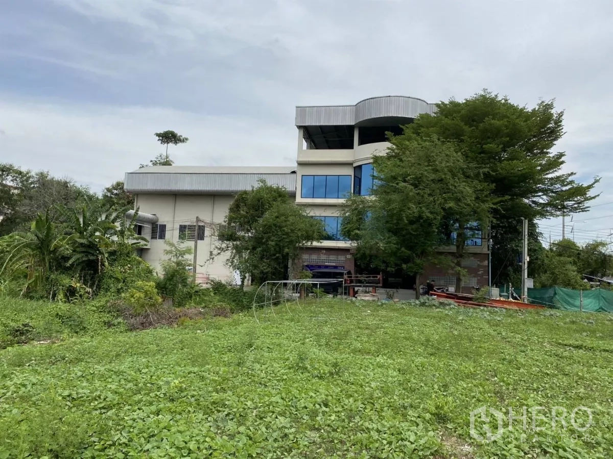 Warehouse for rent or sale in Mueang Nonthaburi, Nonthaburi - Multi-storey office building with curved blue-tinted windows beside a green open field.