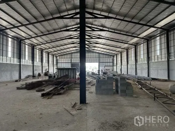 工厂 出租在Phan Thong, Chon Buri - 长条形清爽大跨度仓库空间，中央立柱、混凝土砌块与钢材堆放。