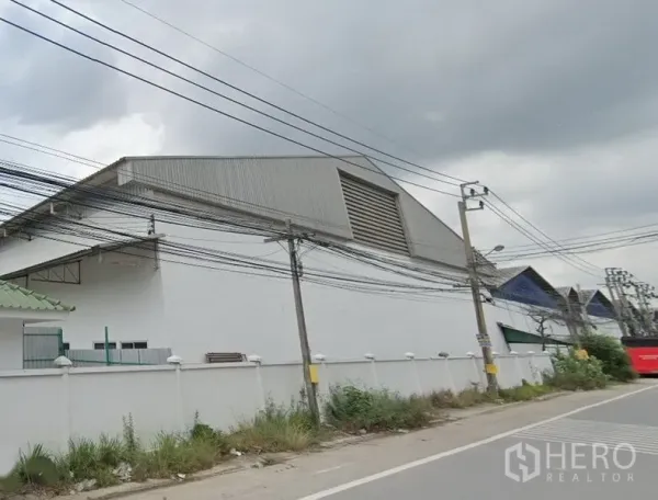 Warehouse for rent in Samut Prakan - Street view of a large white warehouse with high gable metal roof, ventilation louver, perimeter wall and roadside power lines in an industrial area of Bang Sao Thong, Samut Prakan.