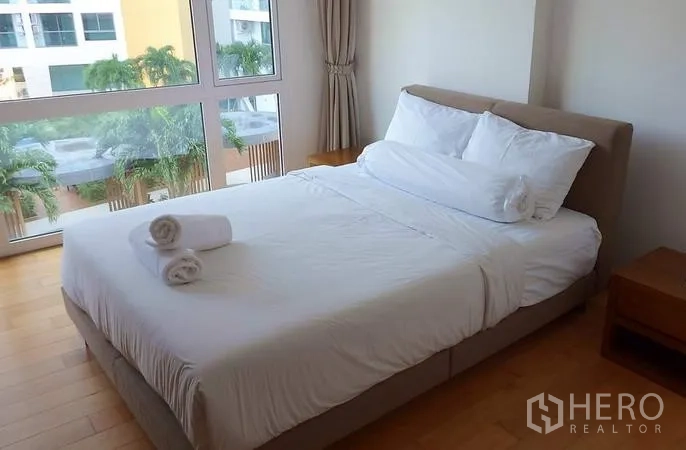 Condominium for sale in Bang Lamung, Chon Buri - Sunlit bedroom with double bed and large windows facing palms and neighboring residences.