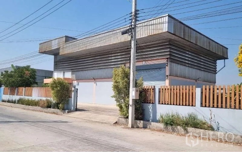 Warehouse for sale in Pathum Thani - Corner view of a metal-clad warehouse with fenced boundary and large roller shutter gate.