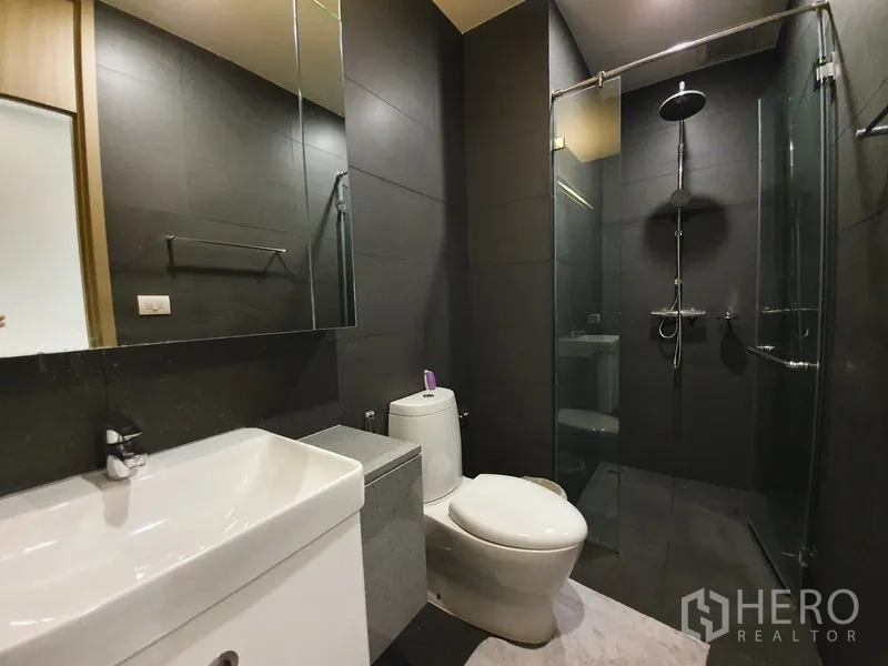 Condominium for rent in Pathum Wan, Bangkok - Sleek bathroom with glass-enclosed rain shower and dark tiled walls.