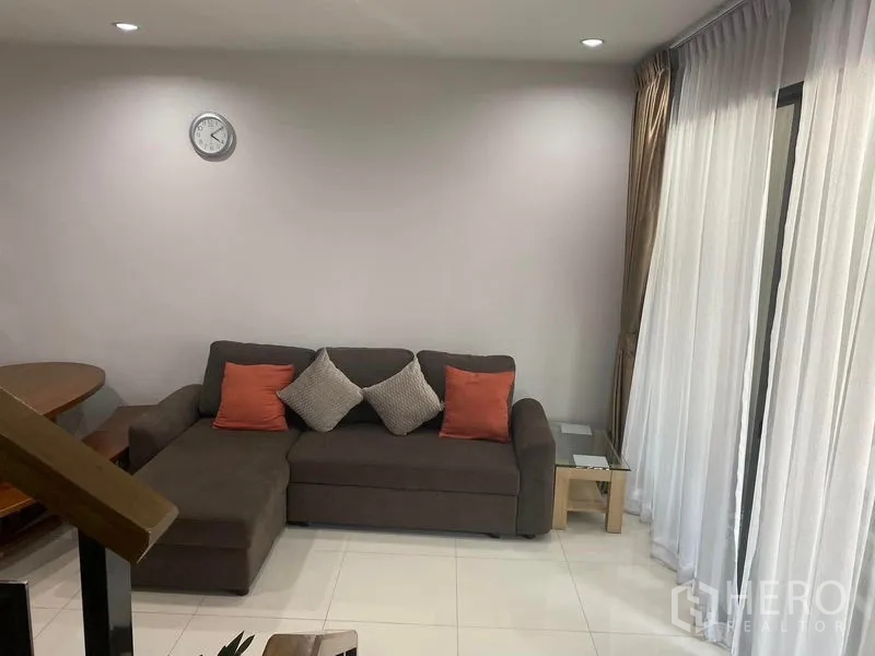 Townhouse for rent or sale in Saphan Sung, Bangkok - Cozy living area with sectional sofa, tiled floor and sliding glass doors with curtains.