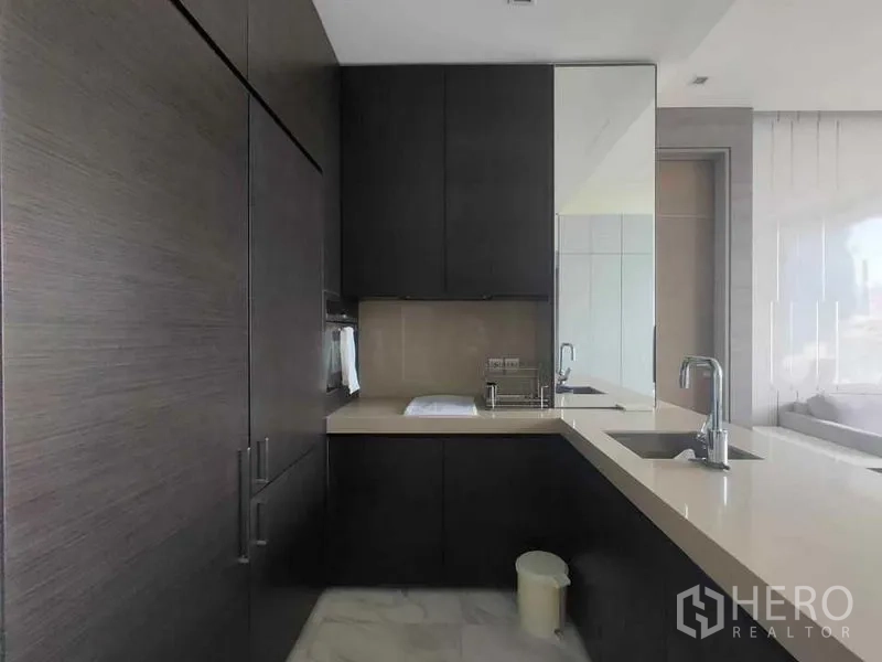 公寓 出租在Bang Rak, Bangkok - L形厨房，深色橱柜、嵌入式烤箱与浅色石材台面及水槽。