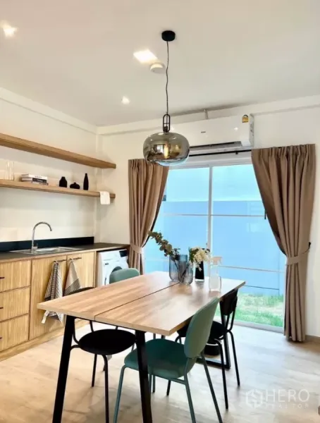 联排别墅 出租在Prawet, Bangkok - 明亮的餐厨区，木质橱柜、吊灯、空调，推拉玻璃门通向小庭院。