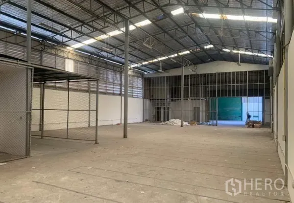 Warehouse for rent in Suan Luang, Bangkok - Spacious warehouse interior with high steel truss roof, skylights, concrete floor, chain-link storage area and open loading access.