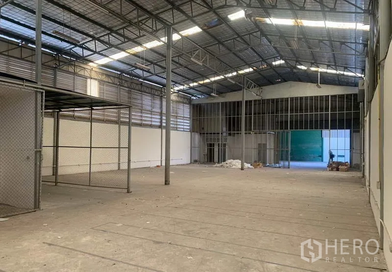 Warehouse for rent in Suan Luang, Bangkok - Spacious warehouse interior with high steel truss roof, skylights, concrete floor, chain-link storage area and open loading access.