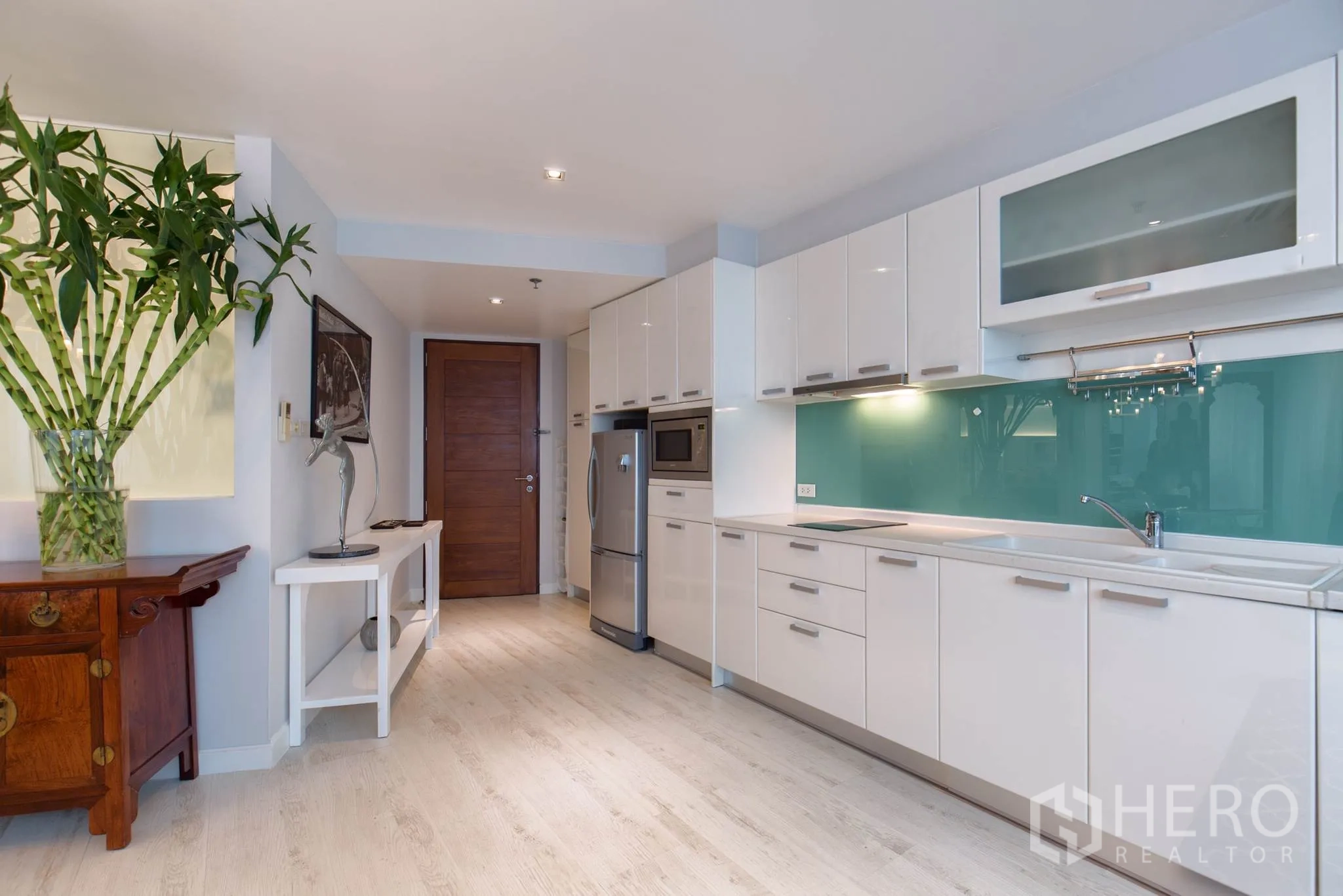 公寓 出租在Pathum Wan, Bangkok - 走廊式厨房，亮白橱柜、不锈钢家电与绿色玻璃背板，浅色木地板。