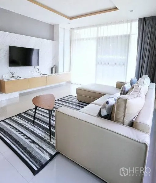 Detached House for rent or sale in Bang Kapi, Bangkok - Sunlit living room with sectional sofa, striped rug, marble TV wall and sheer curtains.
