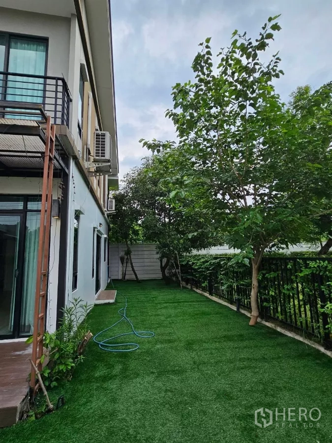 Detached House for rent or sale in Don Mueang, Bangkok - Side yard with a long stretch of green lawn, trees, and black metal fence.