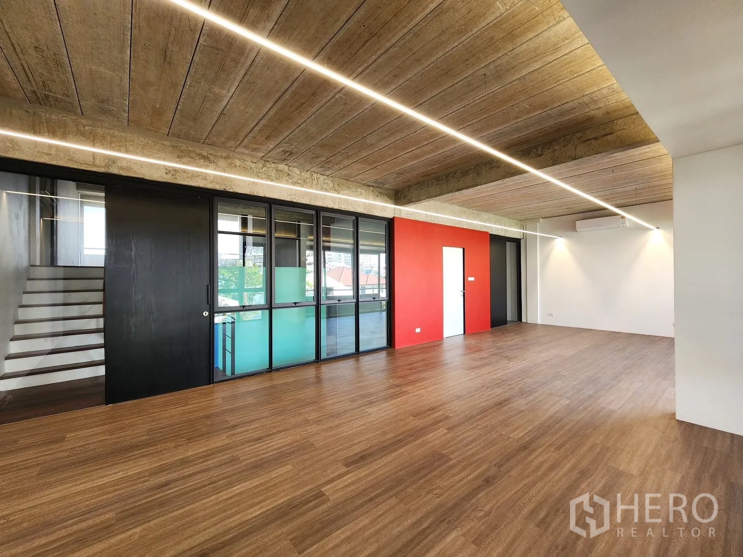 家庭办公室 for rent in Chatuchak, Bangkok - 宽敞工作区，木地板、红色点缀墙，并可见玻璃后方的楼梯。