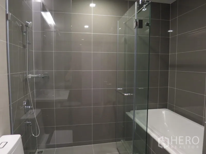 Condominium for rent in Watthana, Bangkok - Modern gray-tiled bathroom with a glass-enclosed shower and a separate bathtub.