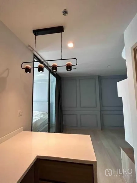 คอนโด ขายในPathum Wan, Bangkok - เคาน์เตอร์แพนทรีพร้อมโคมไฟแขวนสีดำ มองไปยังประตูกระจกบานเลื่อนเข้าสู่ห้องนอนและผนังบิ้วท์ลายคิ้ว