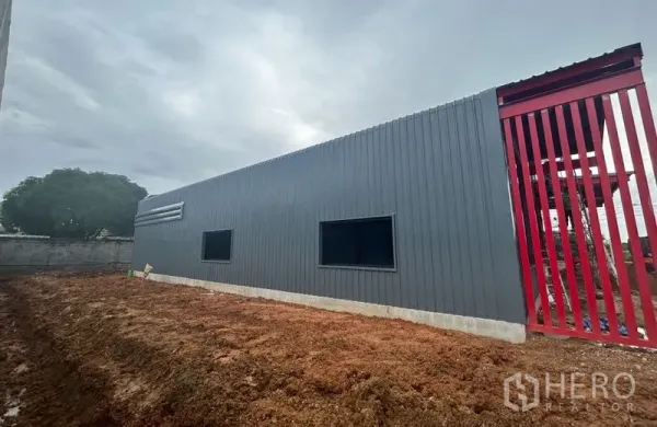 คลังสินค้า ให้เช่าในLam Luk Ka, Pathum Thani - โกดังใหม่ผนังเมทัลชีทสีเทา ประตูกั้นสีแดงและหน้าต่างกรอบดำ ตั้งบนฐานคอนกรีตยกสูง พร้อมลานดินกว้างด้านนอก