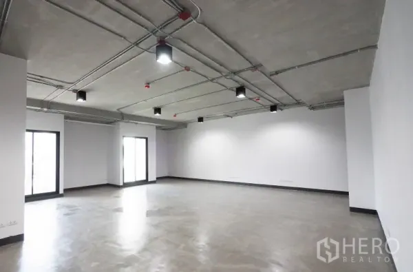 Office Space for rent in Khlong Toei, Bangkok - Spacious corner office with large windows, visible conduits on a concrete ceiling, LED lights, white walls and smooth grey flooring.