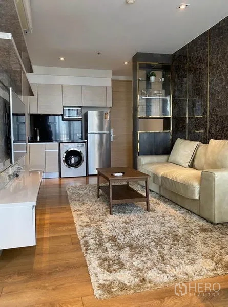 Condominium for rent in Khlong Toei, Bangkok - View of living area toward kitchen with plush rug, leather sofa, and built-in shelving on dark stone panels.