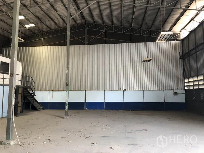 Warehouse for rent in Lam Luk Ka, Pathum Thani - Open storage floor with corrugated metal wall, stairs to mezzanine office, concrete surface and ventilation panels.
