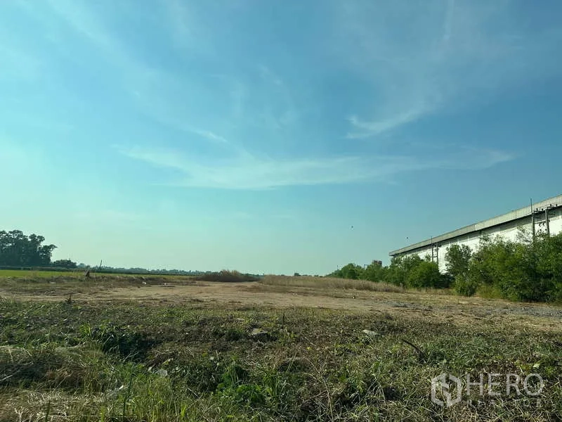 Land for sale in Chachoengsao - Wide, level land ready for development with trees and a long building on the right.