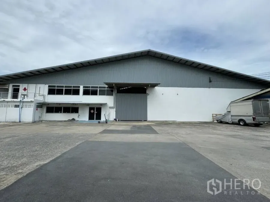 货栈 出租在Mueang Pathum Thani, Pathum Thani - 仓库正面广角，配大型卷闸门和宽敞硬化卡车场地。