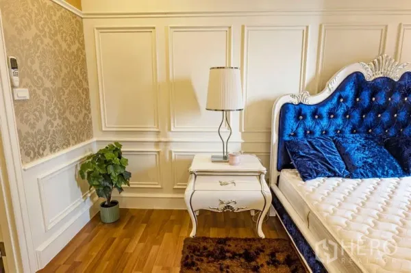 Condominium for rent in Watthana, Bangkok - Bedroom corner with a tufted blue velvet headboard, vintage nightstand and lamp, paneled walls and wood flooring.