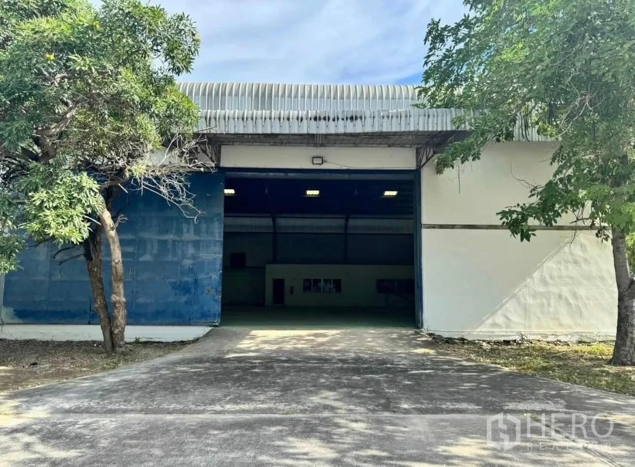Factory for rent in Bang Phli, Samut Prakan - Factory exterior with a large sliding door, concrete driveway, and shade trees.