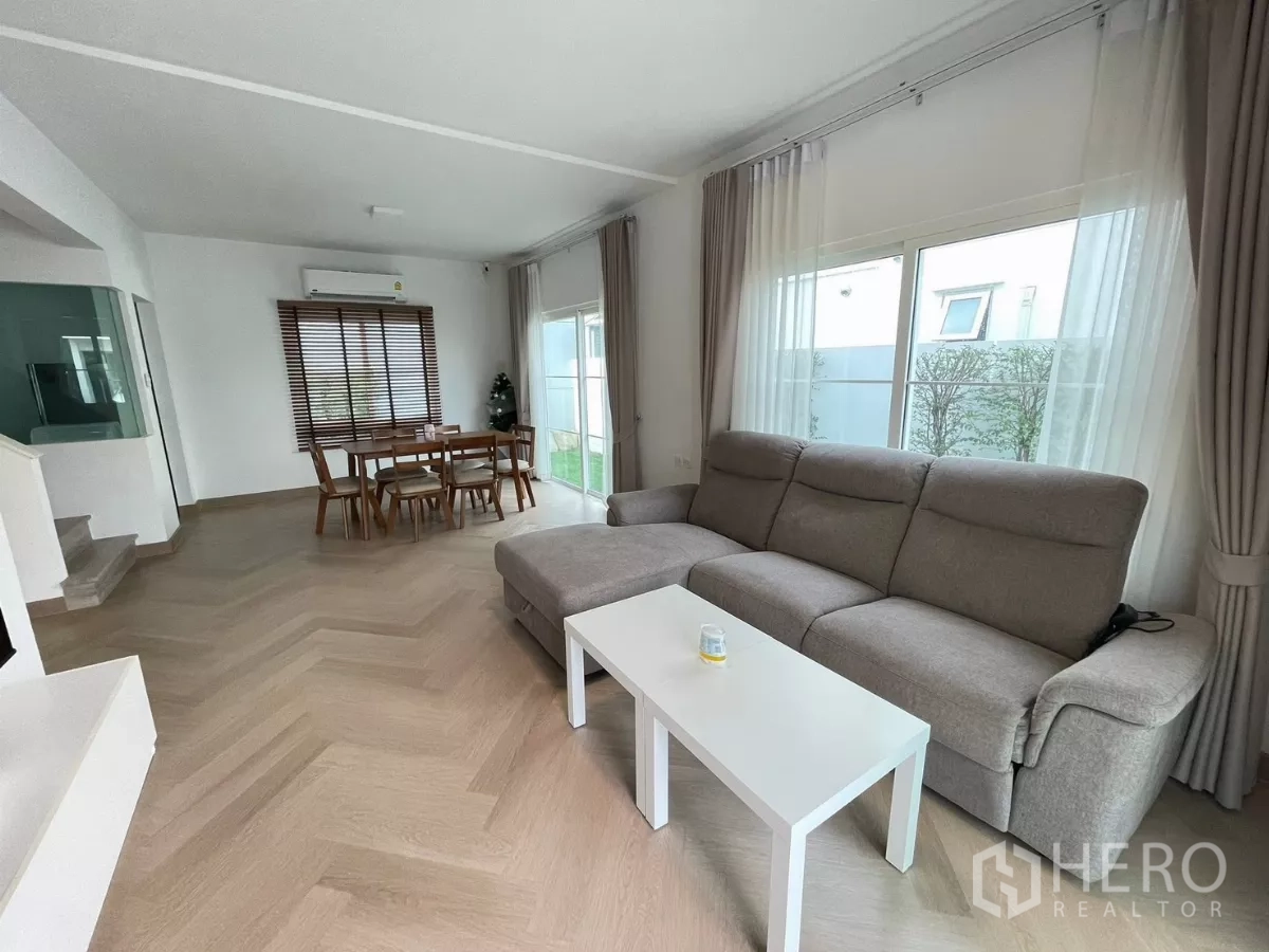Detached House for rent or sale in Bang Phli, Samut Prakan - Bright open-plan living and dining area with an L-shaped sofa, large windows and herringbone wood flooring.