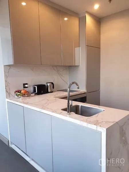 Condominium for rent in Huai Khwang, Bangkok - Bright kitchenette with marble-look countertop, undermount sink, and built-in cabinets with small appliances.