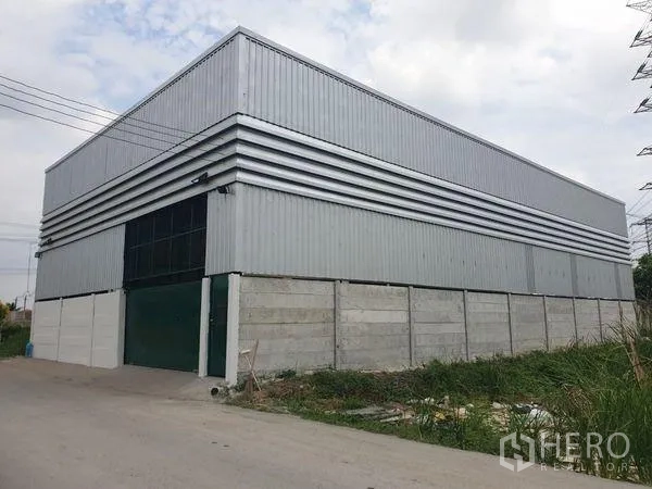 Warehouse for rent in Prawet, Bangkok - Corner view of a metal-clad warehouse with high fence and a wide access road in Prawet.