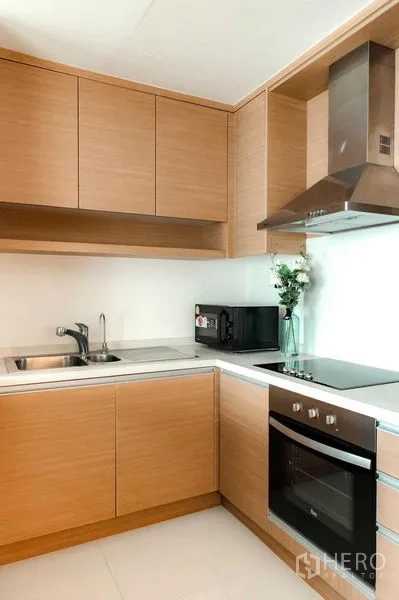 คอนโด ให้เช่าหรือขายในBangkok - ครัวกะทัดรัดมุมแอล เตาอบ เครื่องดูดควัน อ่างล้างจาน และตู้ไม้สีอ่อน