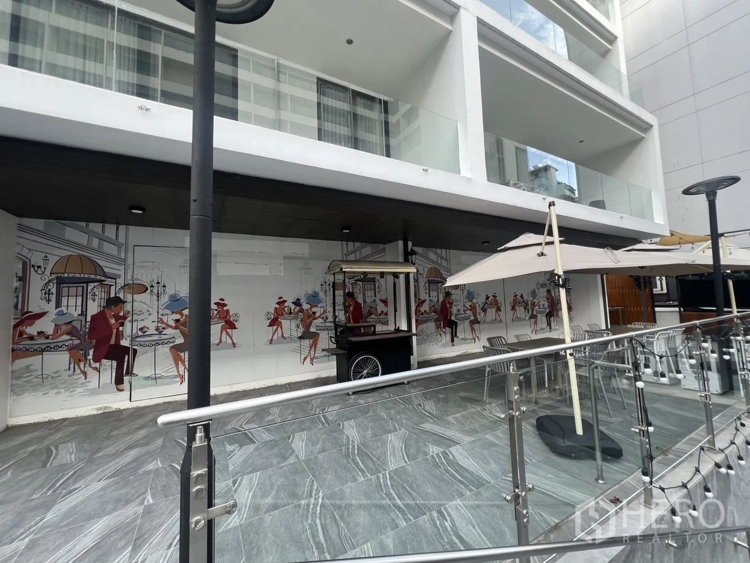 พื้นที่ขายปลีก ให้เช่า - มุมมองด้านหน้าอาคารร้านค้า มีราวกระจก ร่มสนาม และภาพวาดแนวกำแพงตามแนวพลาซ่า