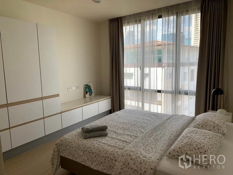 Condominium for rent in Watthana, Bangkok - Master bedroom with large window, built-in wardrobes and a double bed with patterned linens.