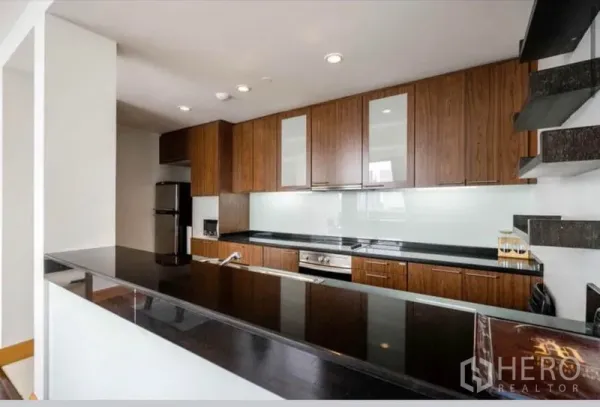 公寓 出租在Sathon, Bangkok - U形现代厨房，木质橱柜、黑色石材台面、玻璃挡板、嵌入式烤箱和不锈钢冰箱。