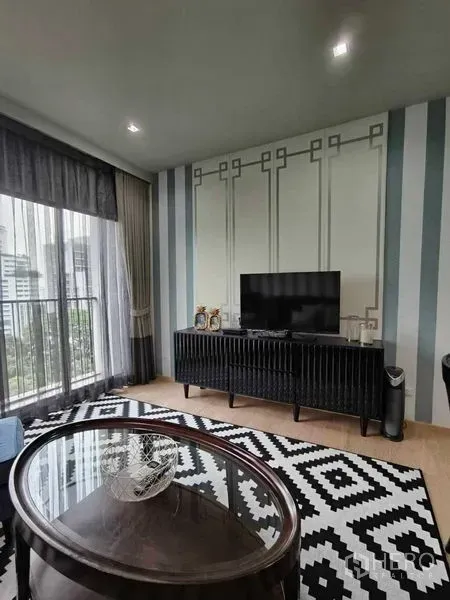 คอนโด ให้เช่าในBangkok - ห้องนั่งเล่นมีตู้วางทีวี พรมลายขาวดำ และหน้าต่างสูงจรดเพดานเปิดออกสู่ระเบียงวิวเมือง