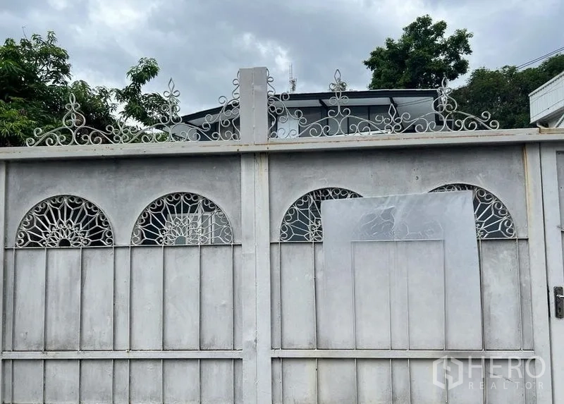 บ้าน ให้เช่าในPhra Khanong, Bangkok - ประตูเหล็กสีขาวลวดลายสวยพร้อมช่องโค้งและต้นไม้ นำสู่บ้านเดี่ยวท่ามกลางท้องฟ้ามีเมฆ