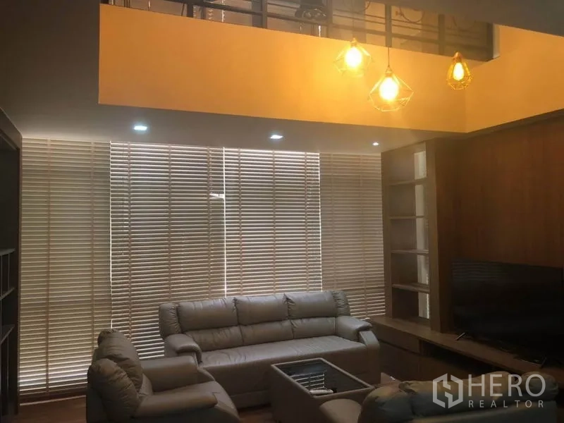 Condominium for sale in Bang Na, Bangkok - Cozy living room with high ceiling, pendant lights, leather sofas, media cabinet and full-width Venetian blinds.