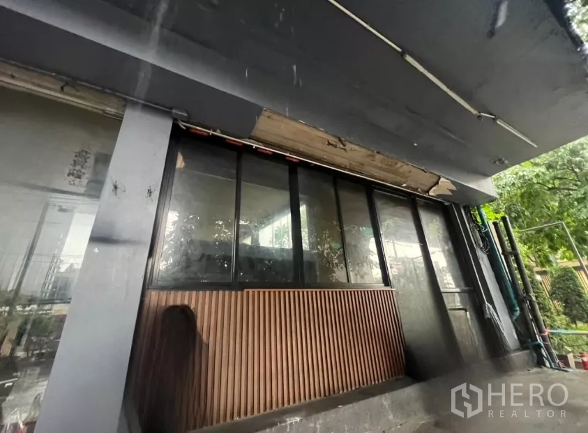อาคารพาณิชย์ ให้เช่าในKhlong Toei, Bangkok - ด้านหน้าร้านระดับถนนมีกันสาดสีดำ กระจกบานใหญ่ และผนังไม้ระแนง ติดพื้นที่สีเขียวริมทาง