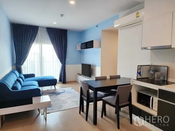 คอนโด ให้เช่า - ห้องนั่งเล่นพร้อมโซฟาตัวยาวสีน้ำเงิน ชุดทานข้าว และครัวเล็กติดระเบียง