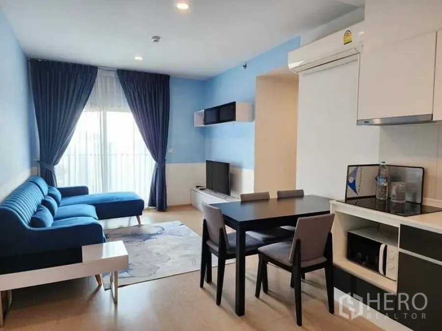 公寓 出租 - 客厅配蓝色转角沙发、餐桌与小厨房，面向阳台。